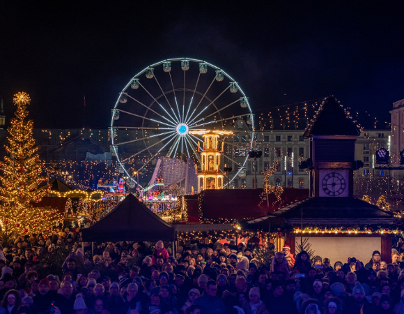 Nocny widok na zatłoczony Jarmark Świąteczny z dużą, jasno oświetloną choinką po lewej i diabelskim młynem w centrum kadru. W dolnej części zdjęcia widoczny jest duży tłum ludzi, a w tle udekorowane światłami budynki.