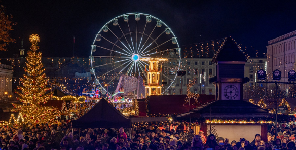 Nocny widok na zatłoczony Jarmark Świąteczny z dużą, jasno oświetloną choinką po lewej i diabelskim młynem w centrum kadru. W dolnej części zdjęcia widoczny jest duży tłum ludzi, a w tle udekorowane światłami budynki.