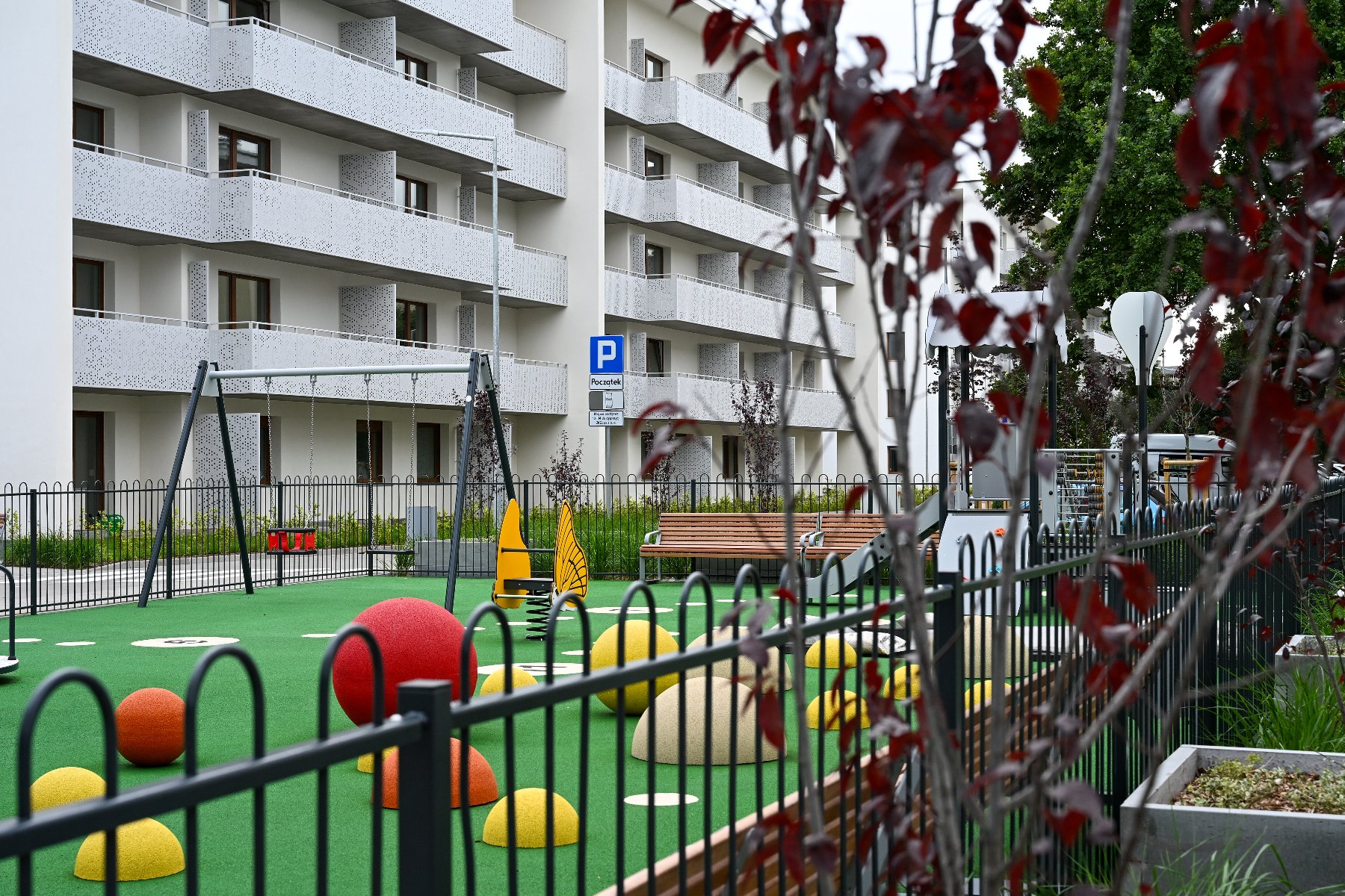 Nowoczesny plac zabaw z zieloną nawierzchnią na osiedlu. Na pierwszym planie czarne metalowe ogrodzenie i gałęzie z czerwonymi liśćmi. Na placu kolorowe półkule, huśtawki oraz żółta zabawka na sprężynie w kształcie motyla. W tle biały wielopiętrowy budynek z licznymi balkonami.