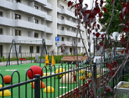 Nowoczesny plac zabaw z zieloną nawierzchnią na osiedlu. Na pierwszym planie czarne metalowe ogrodzenie i gałęzie z czerwonymi liśćmi. Na placu kolorowe półkule, huśtawki oraz żółta zabawka na sprężynie w kształcie motyla. W tle biały wielopiętrowy budynek z licznymi balkonami.