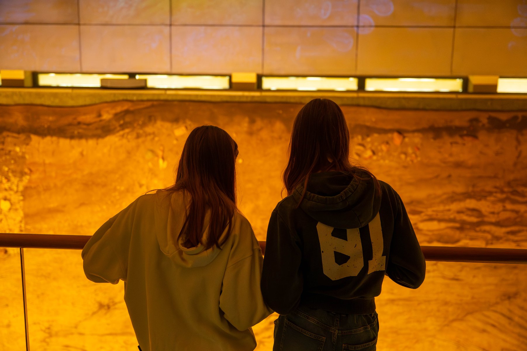 Obraz przedstawia dwie kobiety stojące przy barierce i obserwujące dużą, podświetloną wystawę. Wystawa przypomina stanowisko wykopalisk lub formację geologiczną, oświetloną ciepłym żółto-pomarańczowym światłem, które podkreśla szczegóły ziemi lub warstw skał.