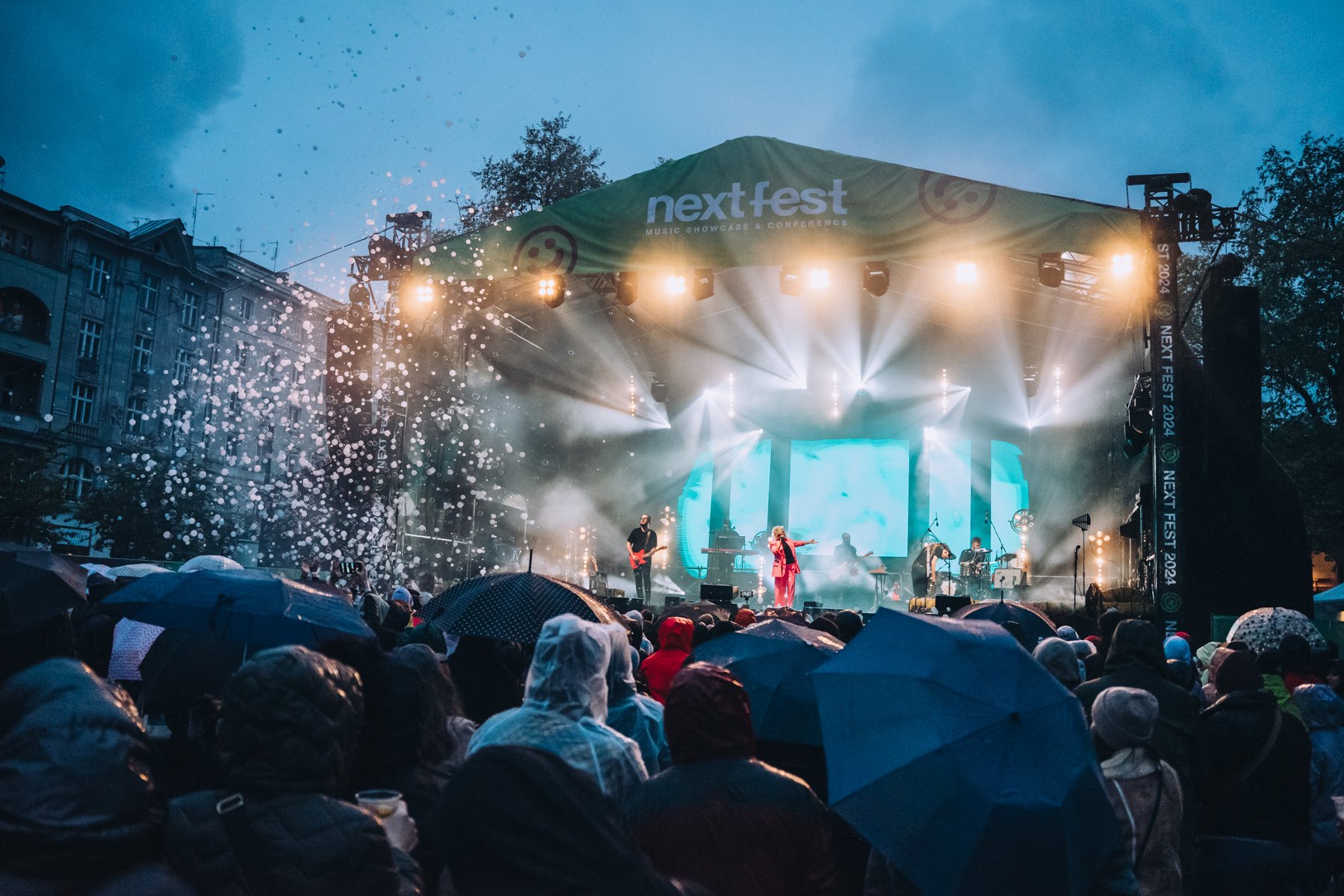 Oświetlona scena z zespołem i wokalistką, przed nią tłum ludzi z parasolami