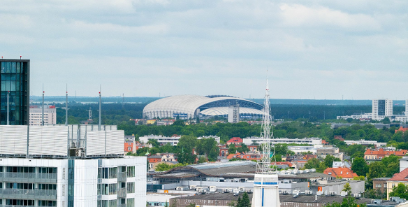 Panorama Poznania z widokiem na Stadion Miejski na horyzoncie. Na pierwszym planie widać nowoczesne budynki mieszkalne i biurowe. Wokół stadionu widać drzewa i zieleń, a w tle rozciąga się las.