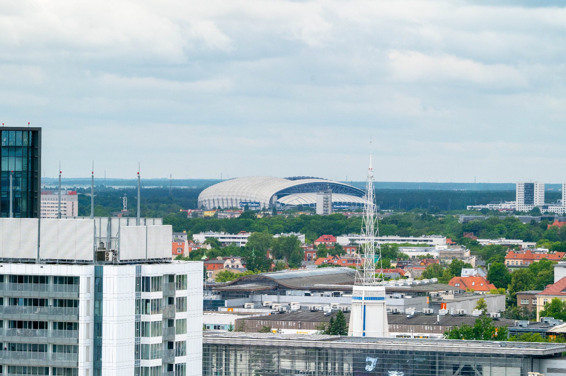Panorama Poznania z widokiem na Stadion Miejski na horyzoncie. Na pierwszym planie widać nowoczesne budynki mieszkalne i biurowe. Wokół stadionu widać drzewa i zieleń, a w tle rozciąga się las.