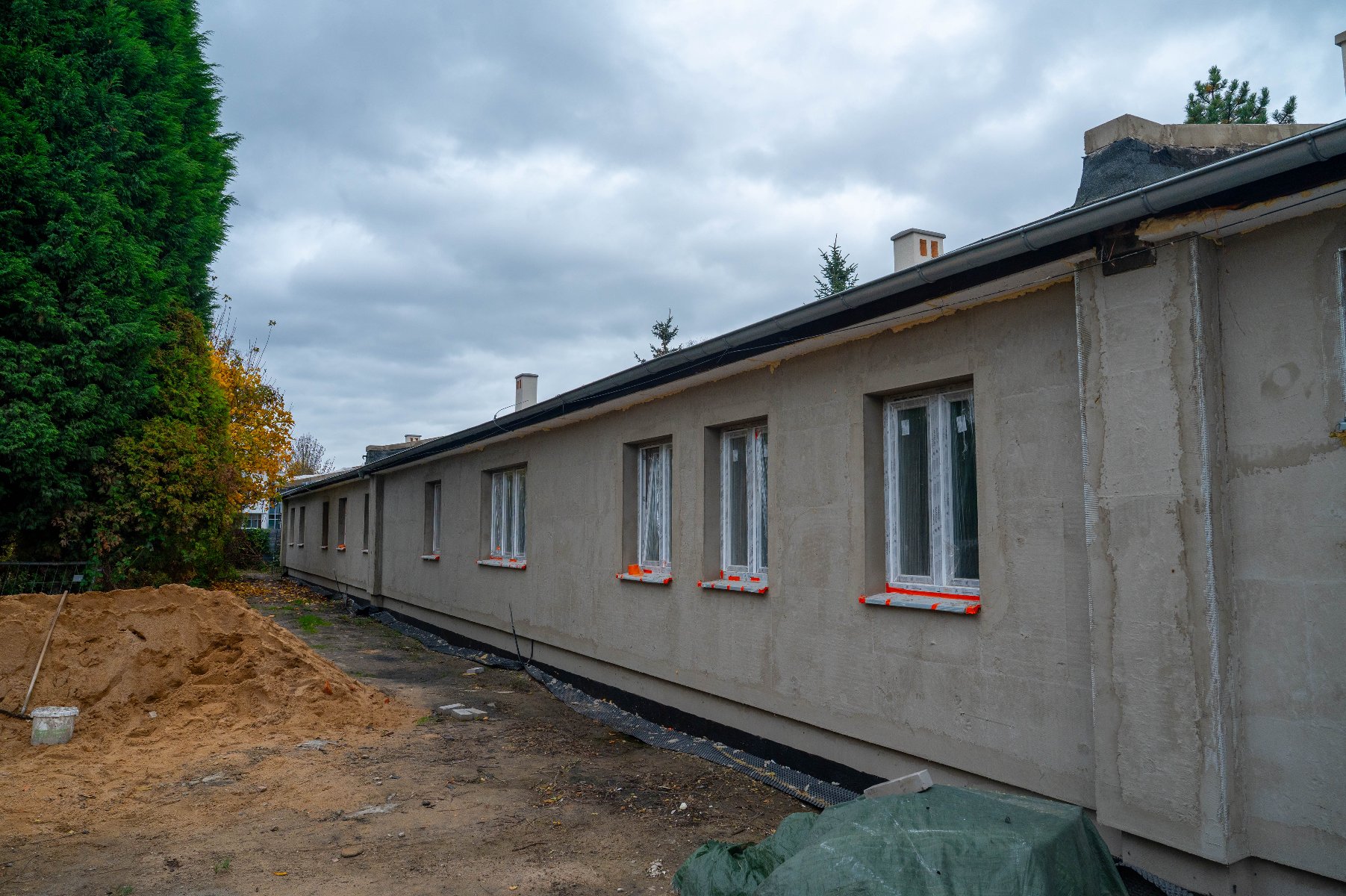 Parterowy budynek w trakcie remontu i docieplania. Ściana pokryta jest szarą zaprawą tynkarską bez koloru, a okna mają widoczne pomarańczowe zabezpieczenia na ramach. Wzdłuż budynku usypana jest duża sterta pomarańczowego piasku i ziemi.