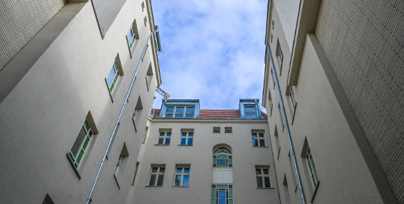 Perspektywiczne ujęcie z dołu przedstawiające wąski dziedziniec otoczony wysokimi, jasnymi ścianami kamienic. W centralnej części widać fasadę z ozdobnymi, witrażowymi oknami o zielonych i niebieskich szybkach. Powyżej znajduje się fragment dachu z czerwoną dachówką i lukarnami, a nad całością góruje błękitne niebo z delikatnymi chmurami.