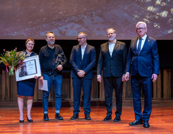 Pięć osób stoi na scenie podczas uroczystości wręczenia nagród. Kobieta po lewej, w niebieskiej sukience, trzyma bukiet kwiatów i oprawiony dyplom. Obok niej stoi mężczyzna w dżinsach i czarnym swetrze, trzymający statuetkę. Pozostali trzej mężczyźni są ubrani w garnitury.