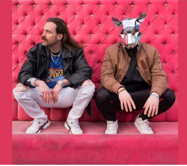 Dwóch mężczyzn kuca, za nimi różowa ściana. Jeden z nich ma na głowie srebrną maskę szczura.