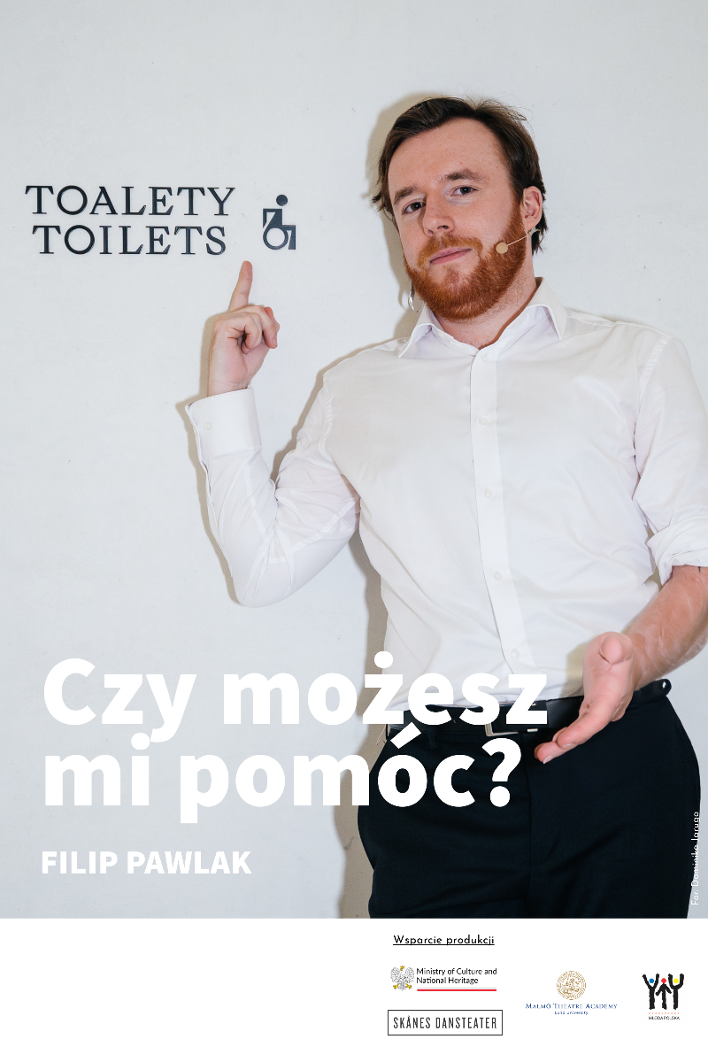 Zdjęcie mężczyzny w białej koszuli na tle białej ściany. Mężczyzna pokazuje palcem napis na ścianie.