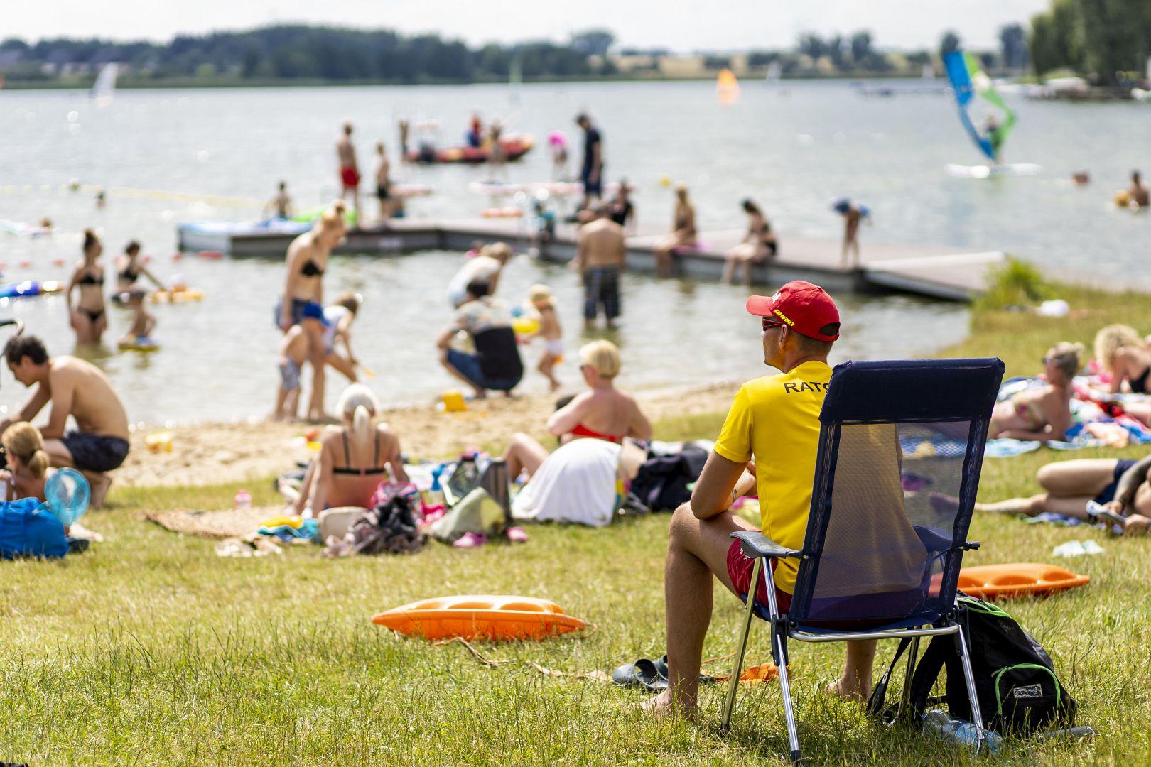plaża nad jeziorem ludzie siedzą na kocach, wielu się kąpie