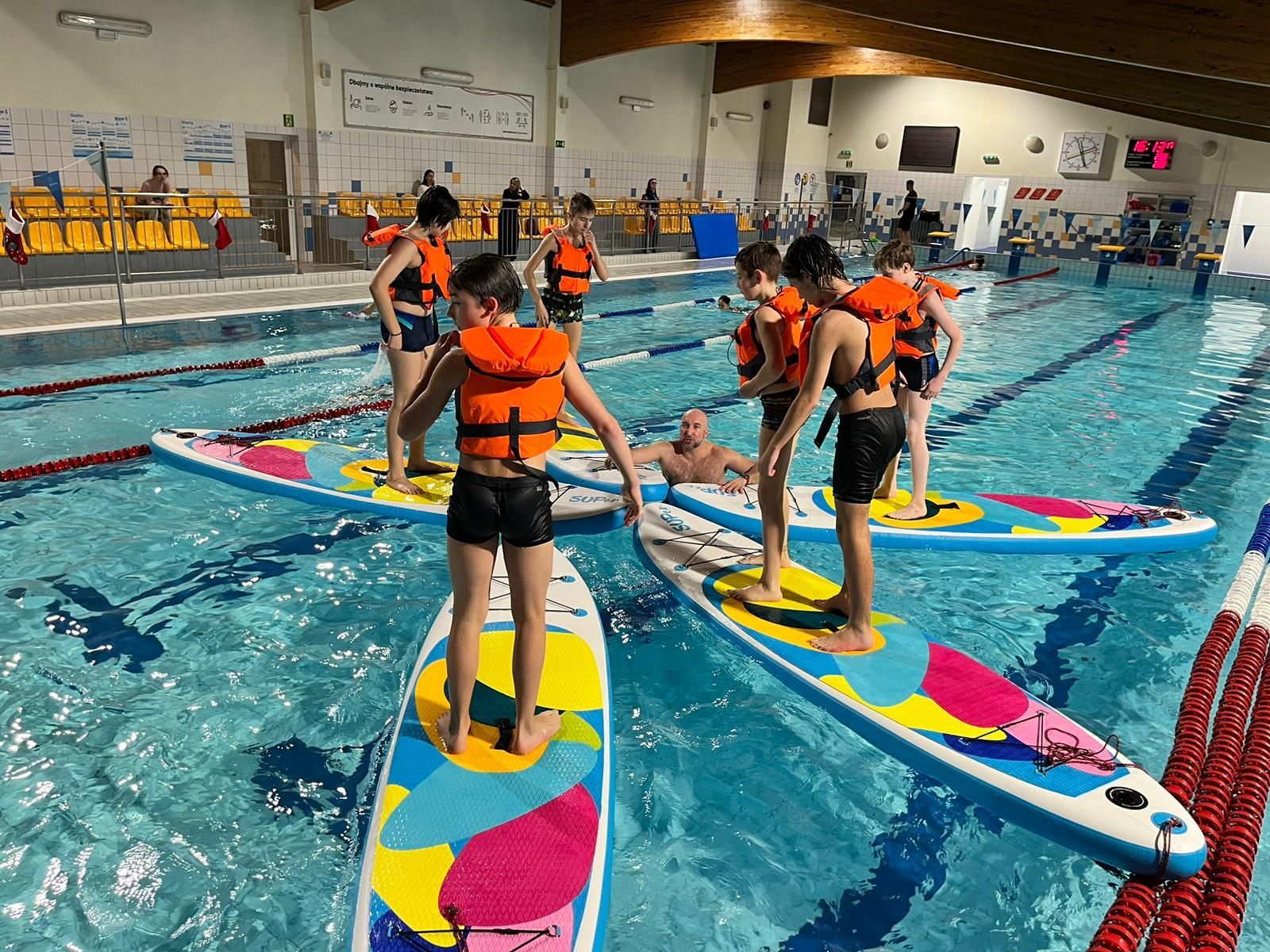 Uczestnicy zajęć stoją w kapokach na deskach SUP. Trener w wodzie.