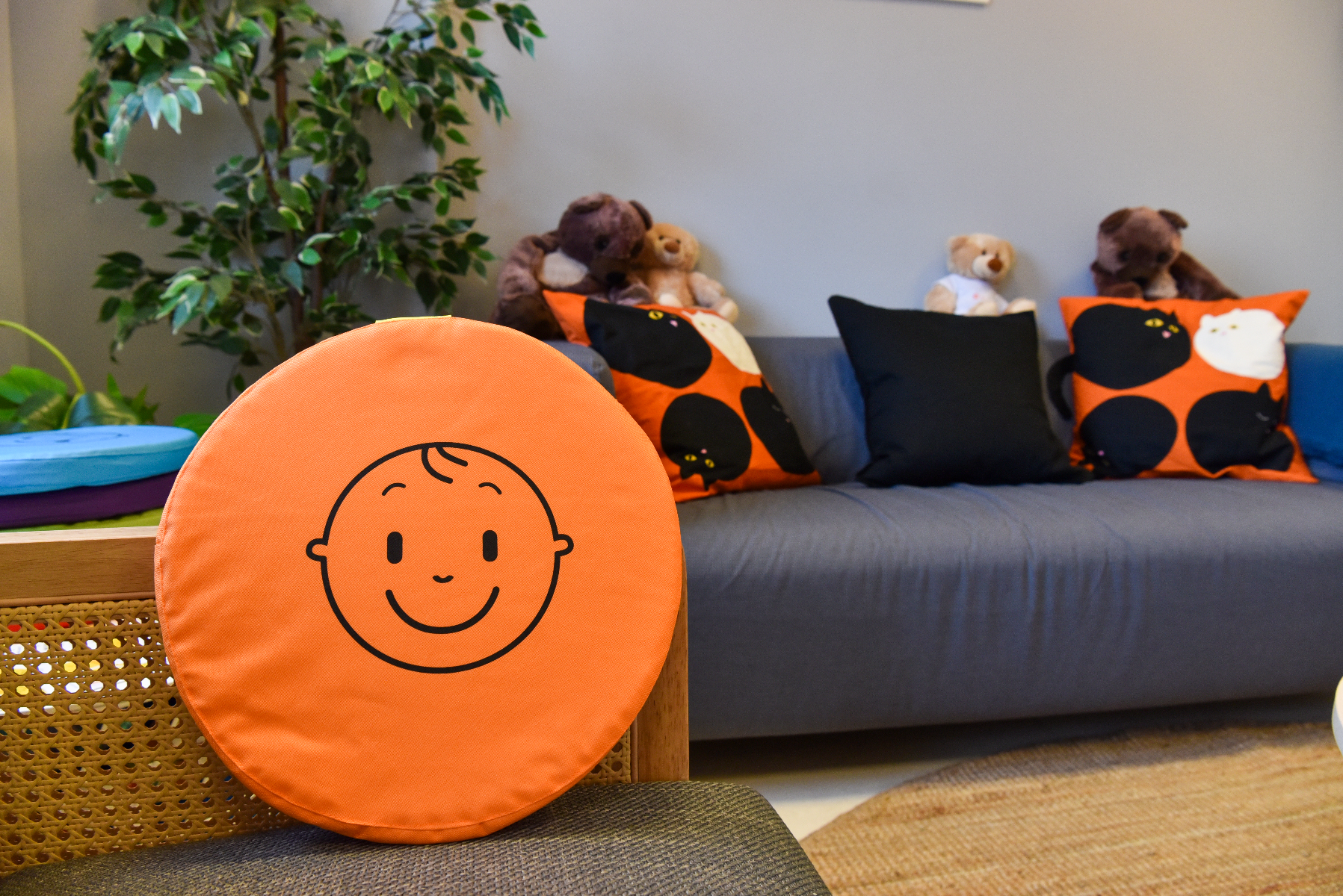Pokój dziecięcy z jasnym i ciepłym wystrojem. Na pierwszym planie znajduje się pomarańczowa poduszka w kształcie okręgu z prostym rysunkiem uśmiechniętej buźki. W tle widać szarą sofę z kilkoma poduszkami ozdobionymi czarnymi i białymi wzorami kota. Na sofie znajdują się także trzy pluszowe misie w różnych kolorach. Z lewej strony pokoju stoi roślina doniczkowa o zielonych liściach.