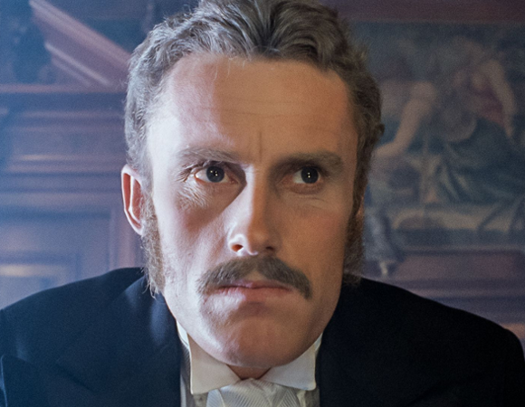 Portretowe ujęcie jasnowłosego mężczyzny z wąsami i baczkami, ubranego w ciemny garnitur z białą koszulą i muszką. Mężczyzna ma surowy wyraz twarzy i patrzy prosto przed siebie. W tle widoczne jest ciemne, drewniane wnętrze z fragmentem dużego obrazu na ścianie.