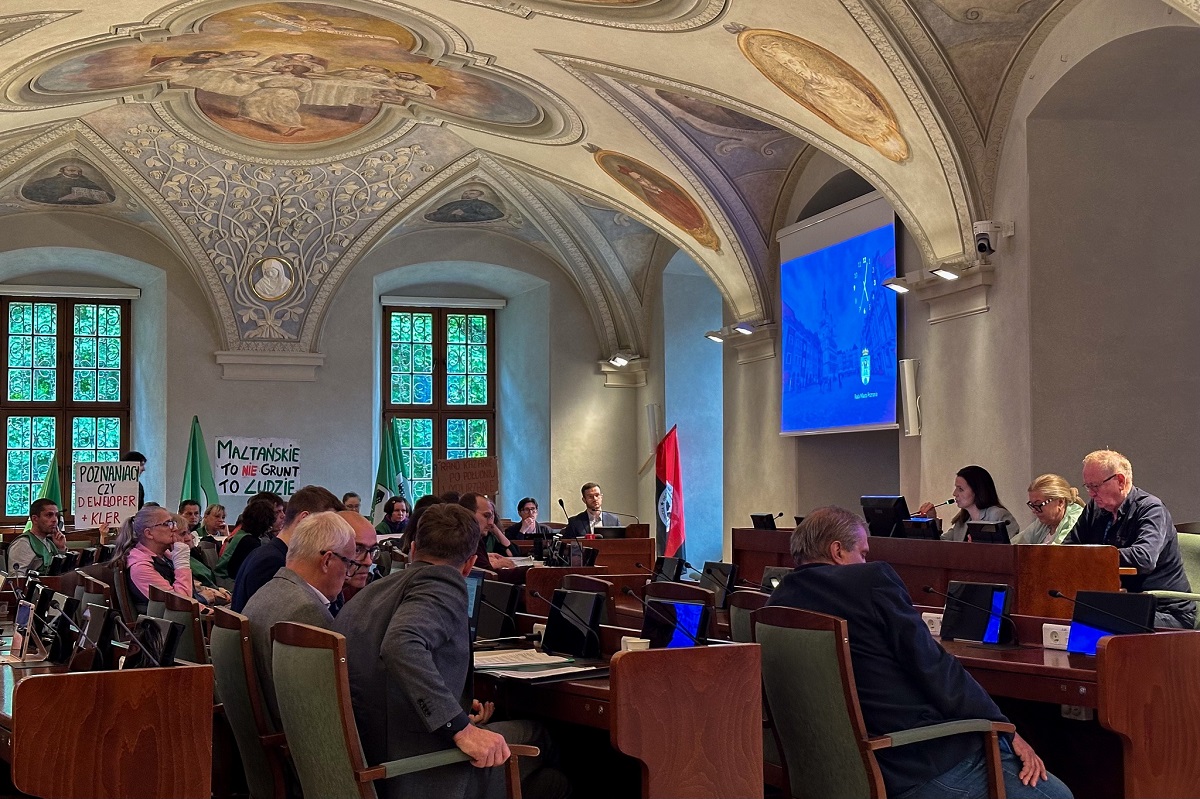 posiedzenie w sali sesyjnej, w rzędach przy pulpitach z monitorami i mikrofonami siedzą ludzie, przed nimi przy stanowisku prezydialnym trzy osoby, w tele siedzą osoby z flagami i transparentami