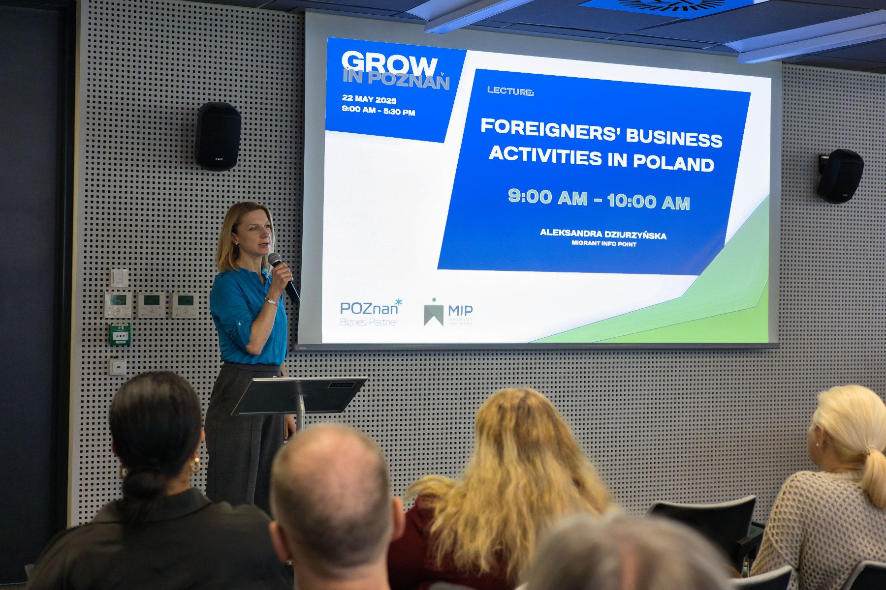 Prelegentka przeprowadza szkolenie z podatków podczas konferencji. W tle wyświetlana grafika z tytułem szkolenia w kolorach granatowym, białym i limonkowym. Szkolenie odbywa się w Plus Jeden. Uczestnicy siedzą i słuchają.