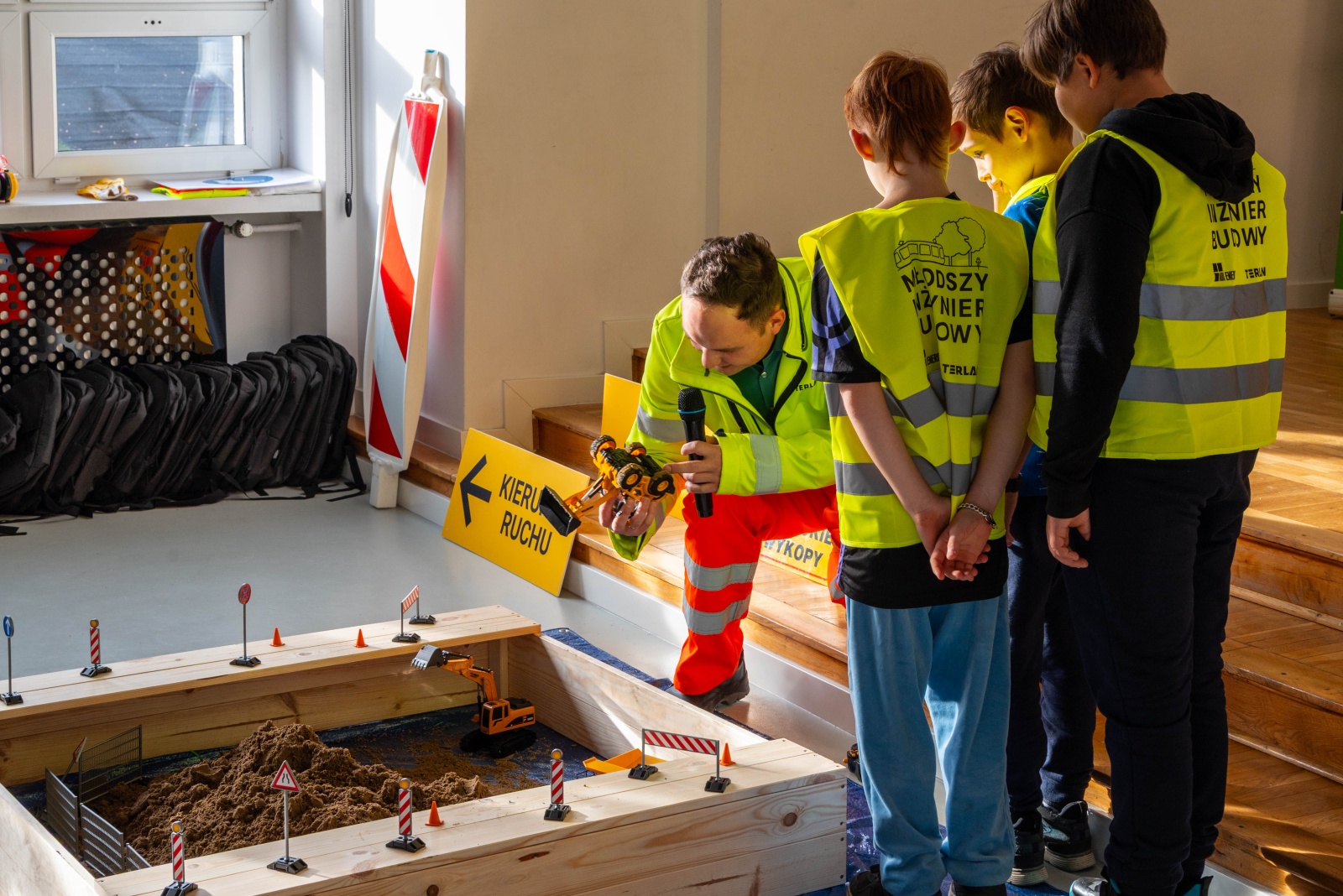 Prowadzący zajęcia mężczyzna nachyla się nad makietą placu budowy. Trzyma w ręku miniaturową koparke i przez mikrofon opowiada. Obok niefo stoji kilkoro dzieci ubranych w odblaskowe kamizelki z napisem: młodszy inżynier budowy.
