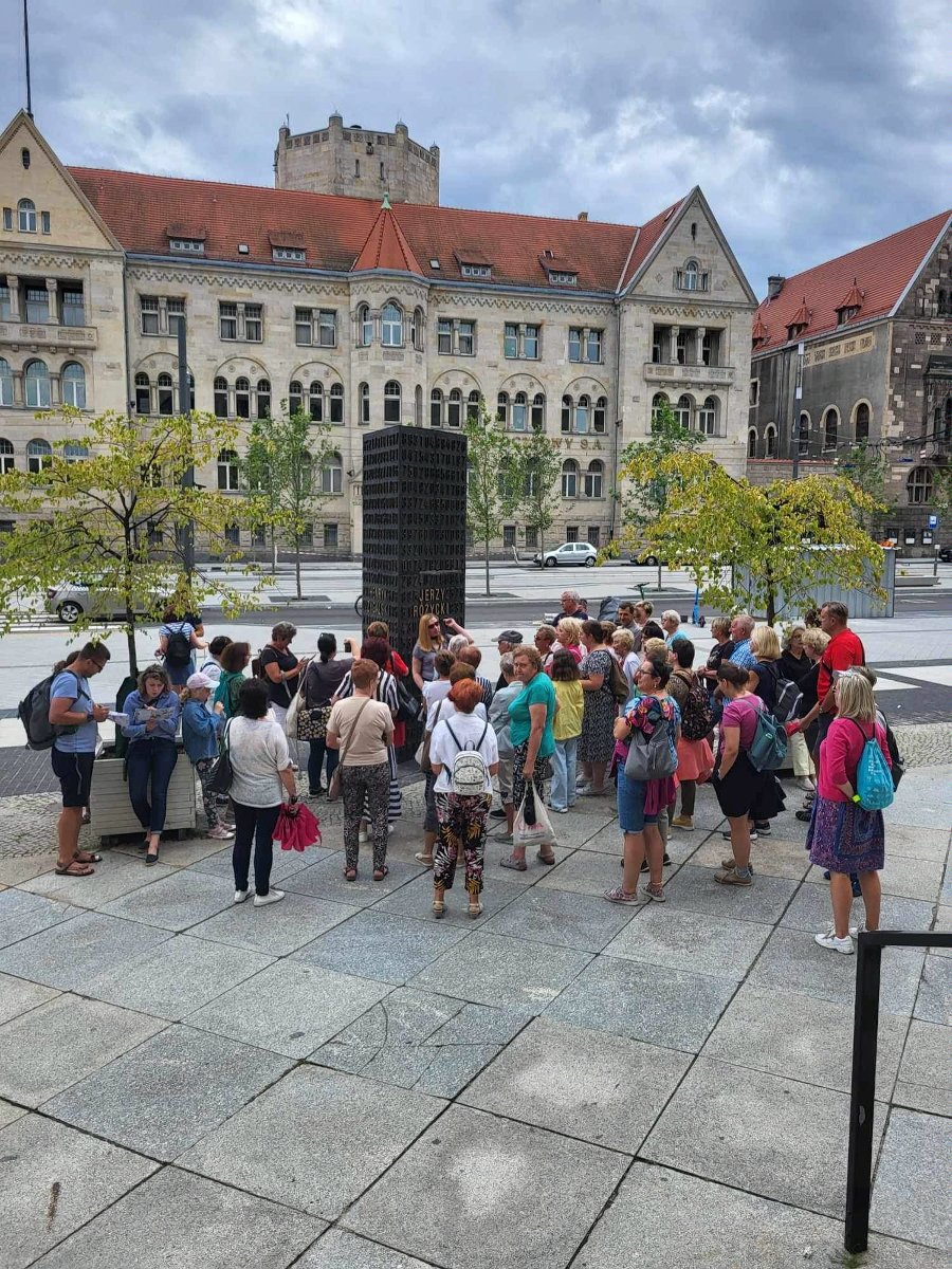 Grupa wycieczkowa stoi przy pomniku kryptologów. Pomnik składa się z prostokątnej bryły z wyrytymi cyframi. Wśród nich są nazwiska kryptologów. Wycieczkowicze są w różnym wieku, ale wspólnie słuchają przewodniczki. W tle budynek Banku Pocztowego na ul. Święty Marcin.