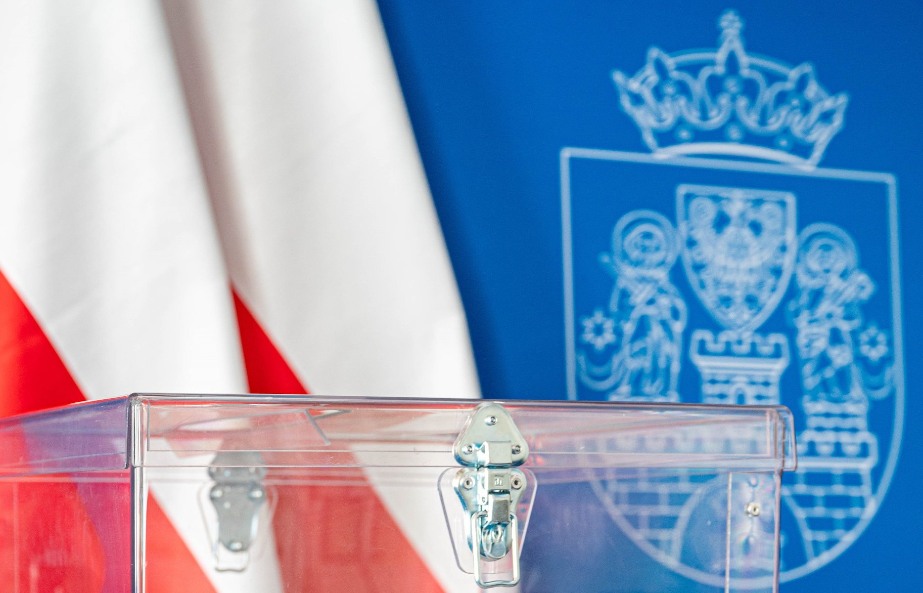 przeźroczysta urna wyborcza w tle dwie flagi biało-czerwone i biały herb miasta na niebieskim tle.