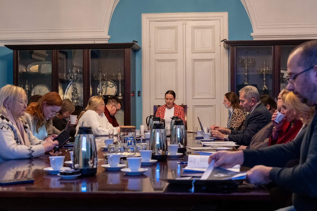 Przy stole konferencyjnym obraduje około kilkanaścioro osób, kobiet i mężczyzn w różnym wieku