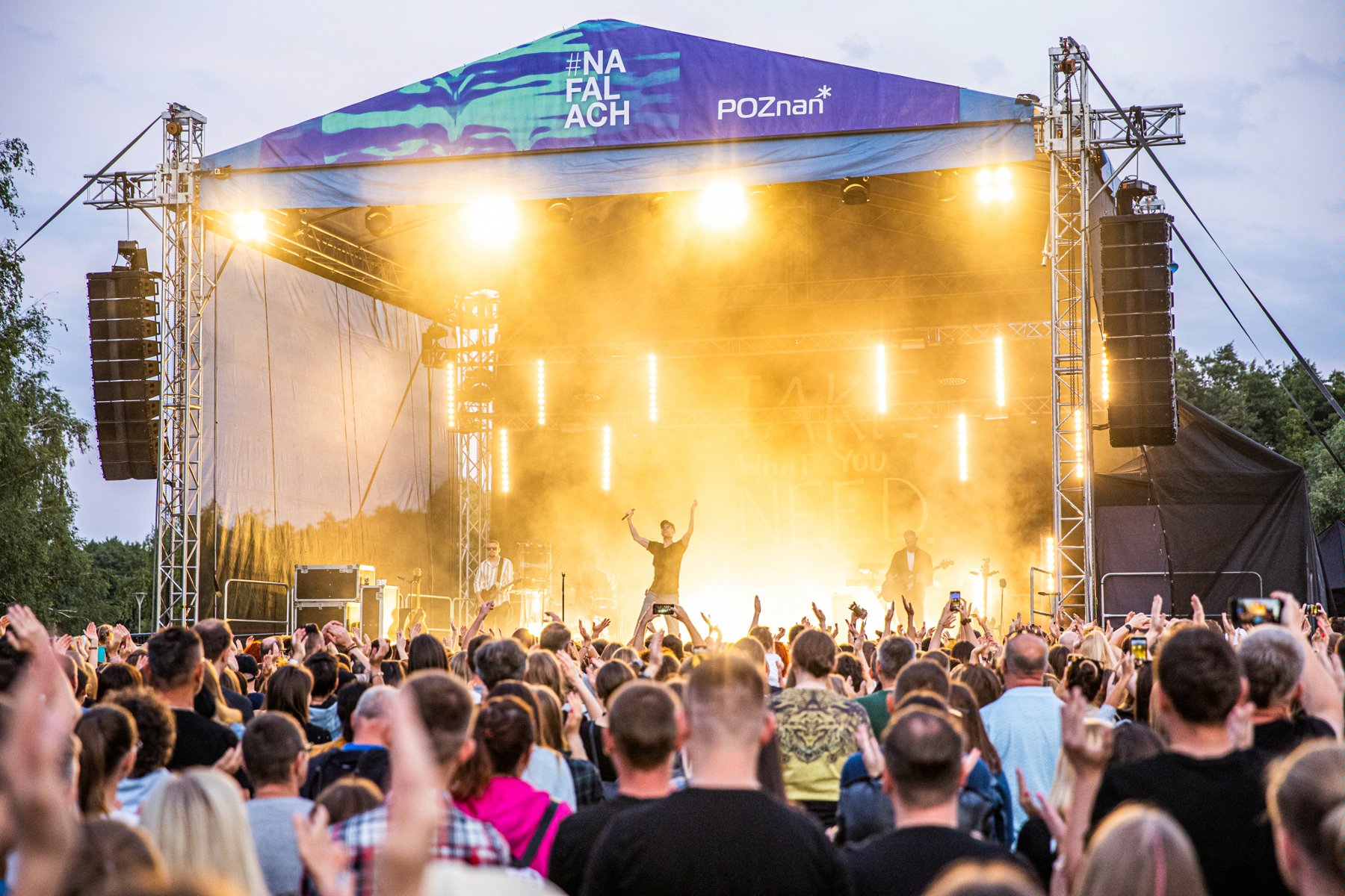 Publiczność stoi i pod sceną i bawi się do muzyki. Na scenie stoi mężczyzna z mikrofonem w ręce i unosi ręce do góry. Scena jest oświetlona na żółto. W tle drzewa.