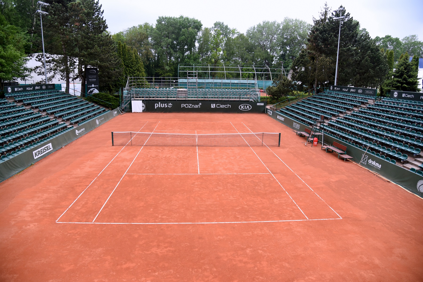 Puste korty tenisowe z czerwoną nawierzchnią otoczone zielonymi trybunami. Na barierkach wokół kortu widoczne są loga sponsorów, m.in.: "KREISEL", "plus", "POZnań", "Ciech", "KIA" i "drutex". Kort otaczają drzewa i zieleń. W głębi widoczna jest konstrukcja, prawdopodobnie trybuna lub platforma widokowa.