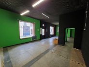 Puste wnętrze lokalu użytkowego o jasnej, betonowej podłodze i czarnym suficie z fakturowanej płyty. Ściana po lewej jest pomalowana na intensywnie zielony kolor. Na tej ścianie znajduje się duże okno, w którym odbija się na szybie tekst "Lokal do wynajęcia. Aukcja".