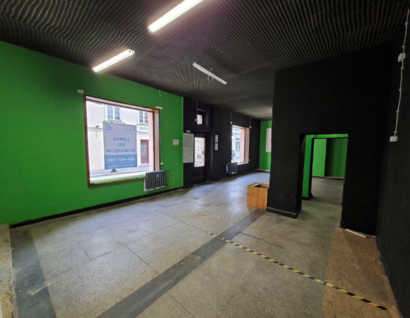 Puste wnętrze lokalu użytkowego o jasnej, betonowej podłodze i czarnym suficie z fakturowanej płyty. Ściana po lewej jest pomalowana na intensywnie zielony kolor. Na tej ścianie znajduje się duże okno, w którym odbija się na szybie tekst "Lokal do wynajęcia. Aukcja".