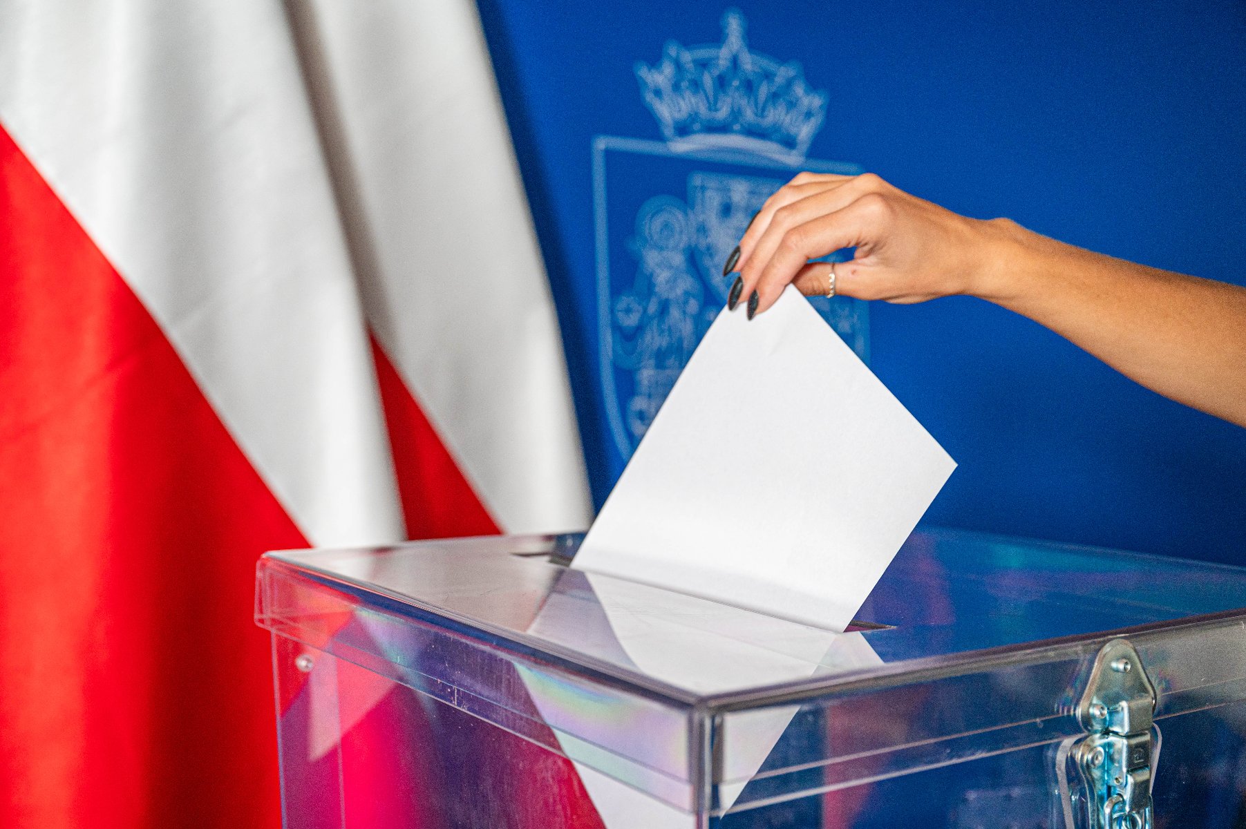 Ręka osoby wrzuca głos wyborczy do przeźroczystej urny wyborczej. W tle biało-czerwone flagi i herb Miasta Poznania na niebieskim tle.
