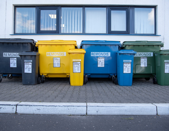 Rząd czarnych, żółtych, niebieskich i zielonych dużych i małych kontenerów na śmieci, ustawionych przed białą fasadą budynku z oknami w ciemnych ramach.