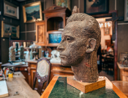 Rzeźba głowy mężczyzny wykonana z ciemnego, chropowatego materiału, ustawiona na drewnianej podstawie i zielonym postumencie. Popiersie ukazane jest z profilu. W tle widoczne jest bogato wyposażone wnętrze gabinetu lub antykwariatu: rzeźbione szafy, liczne obrazy w złotych ramach na ścianach, krzesło z ozdobną tapicerką oraz biurko z akcesoriami piśmienniczymi