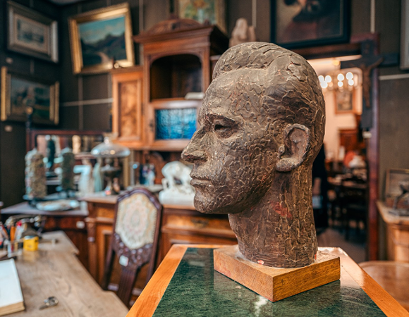 Rzeźba głowy mężczyzny wykonana z ciemnego, chropowatego materiału, ustawiona na drewnianej podstawie i zielonym postumencie. Popiersie ukazane jest z profilu. W tle widoczne jest bogato wyposażone wnętrze gabinetu lub antykwariatu: rzeźbione szafy, liczne obrazy w złotych ramach na ścianach, krzesło z ozdobną tapicerką oraz biurko z akcesoriami piśmienniczymi