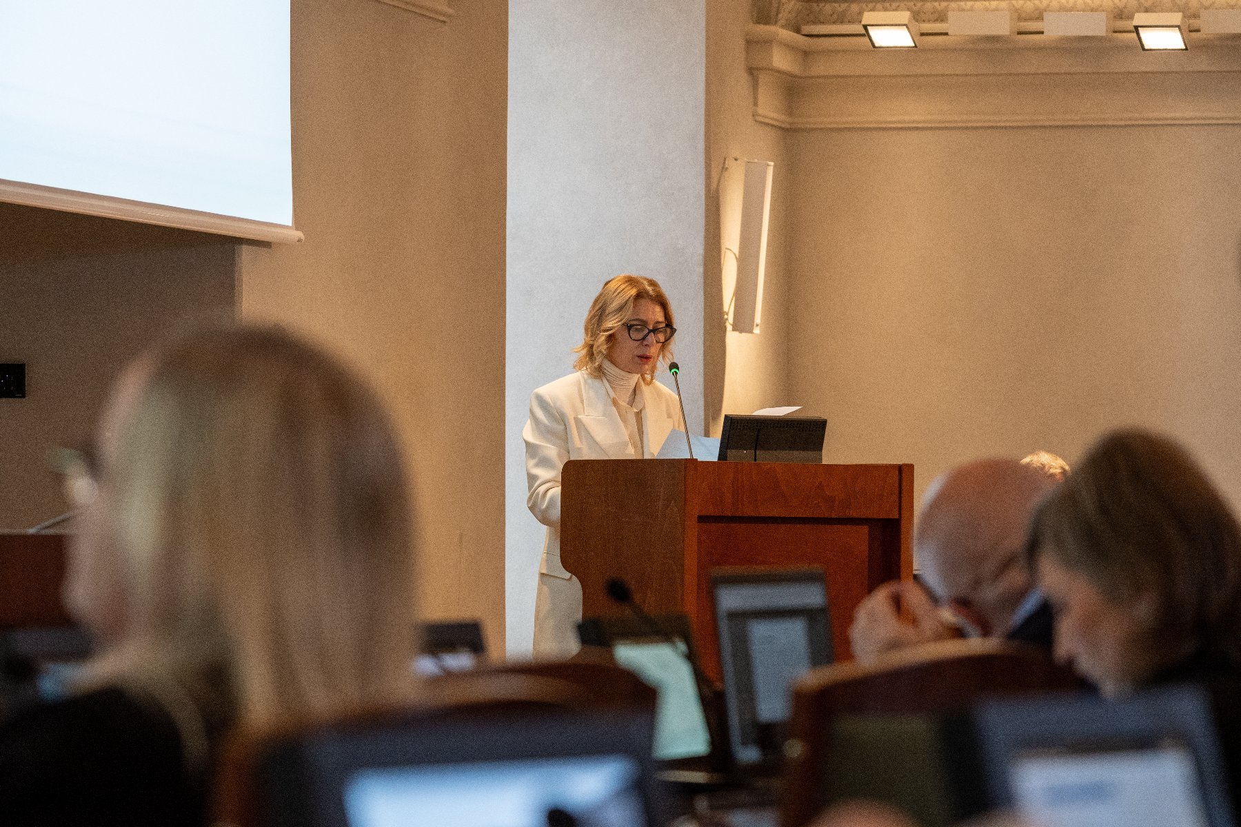 Sala sesyjna, przy mównicy kobieta w okularach, ubrana na biało