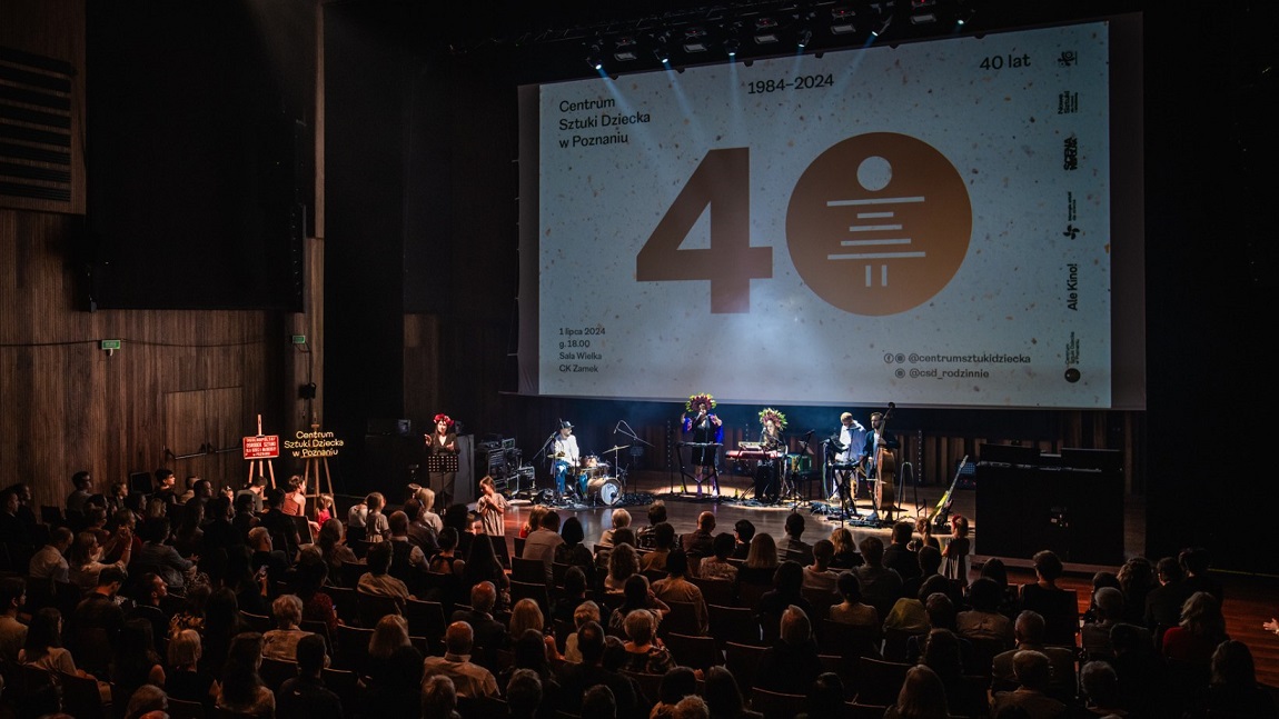 scena, na której jest duży ekran świetlny, a na nim wyświetlona duża liczba 40, pod ekranem zespół muzyczny, na widowni sposo osób