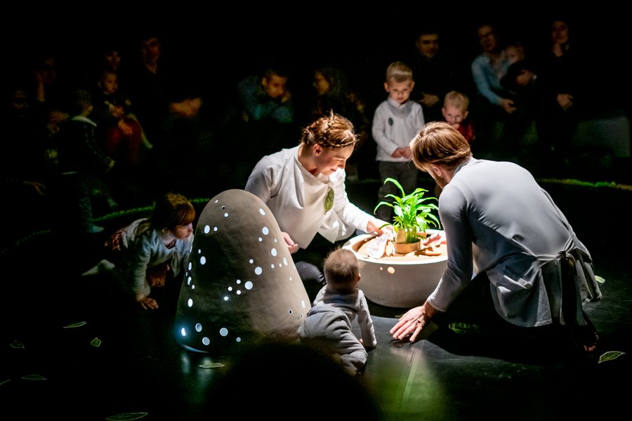 Scena, na której występuje dwoje tancerzy. Pochylają się oni nad kwiatkiem. Dookoła zainteresowne dzieci.