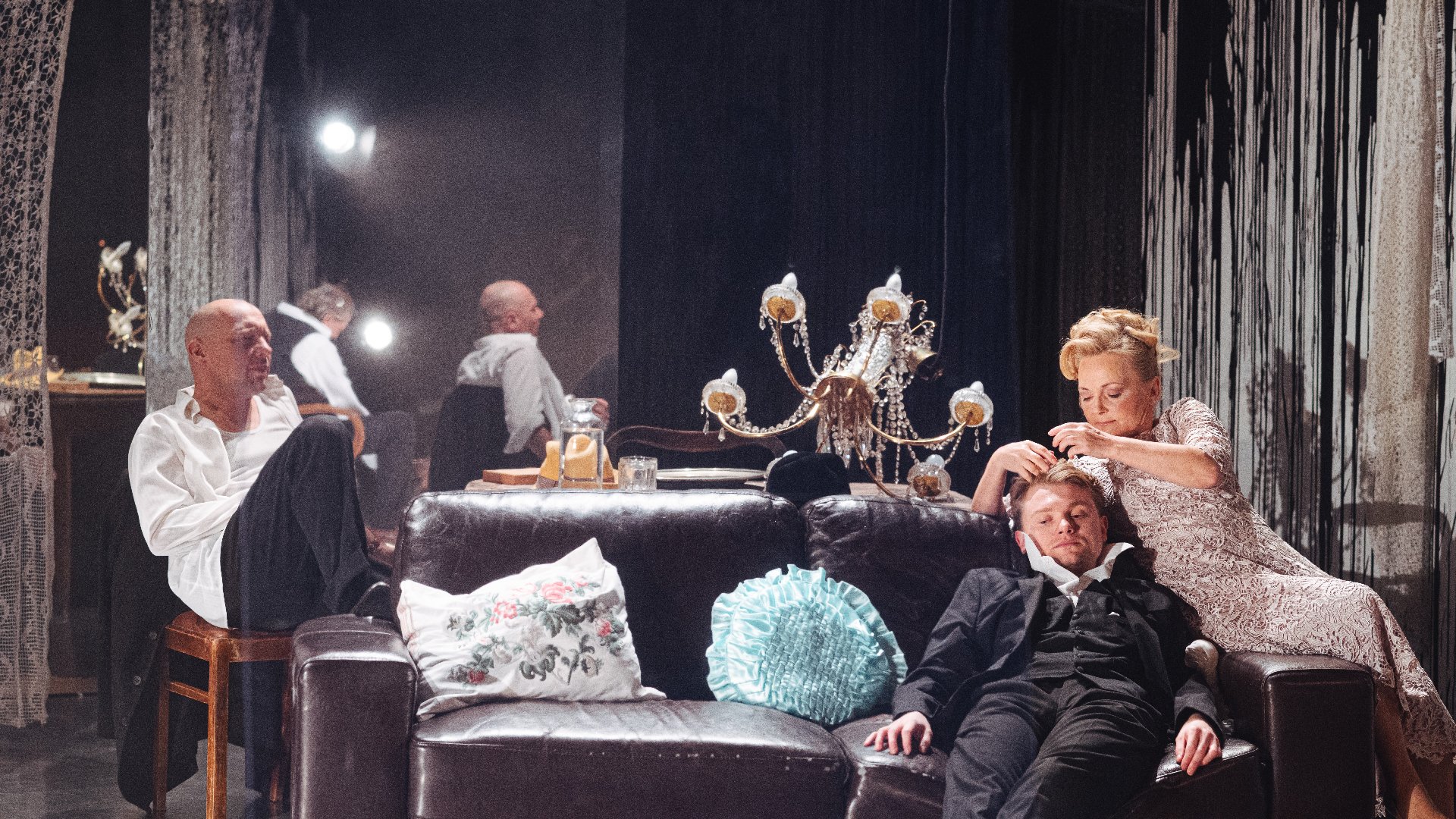 Scena teatralna. W półmroku eleganckiego wnętrza mężczyzna siedzi na stołku przy stole, a na kanapie leży młody mężczyzna w czarnym garniturze, któremu kobieta czesze włosy. Na pierwszym planie sofa z dekoracyjnymi poduszkami i żyrandol; w tle lustra i światła sceniczne.