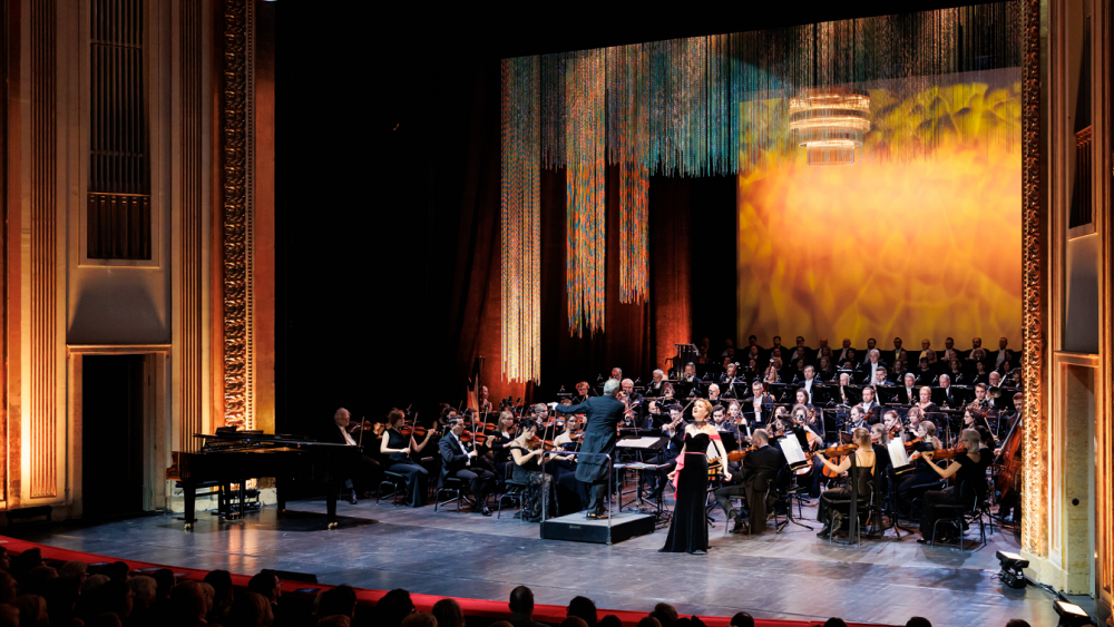 Scena w Teatrze Wielkim jest wypełniona orkiestrą, ekran za orkiestrą ma złotą barwę. Z przodu sceny stoi, tyłem, dyrygent, oraz przodem, śpiewaczka w czarnej sukni.