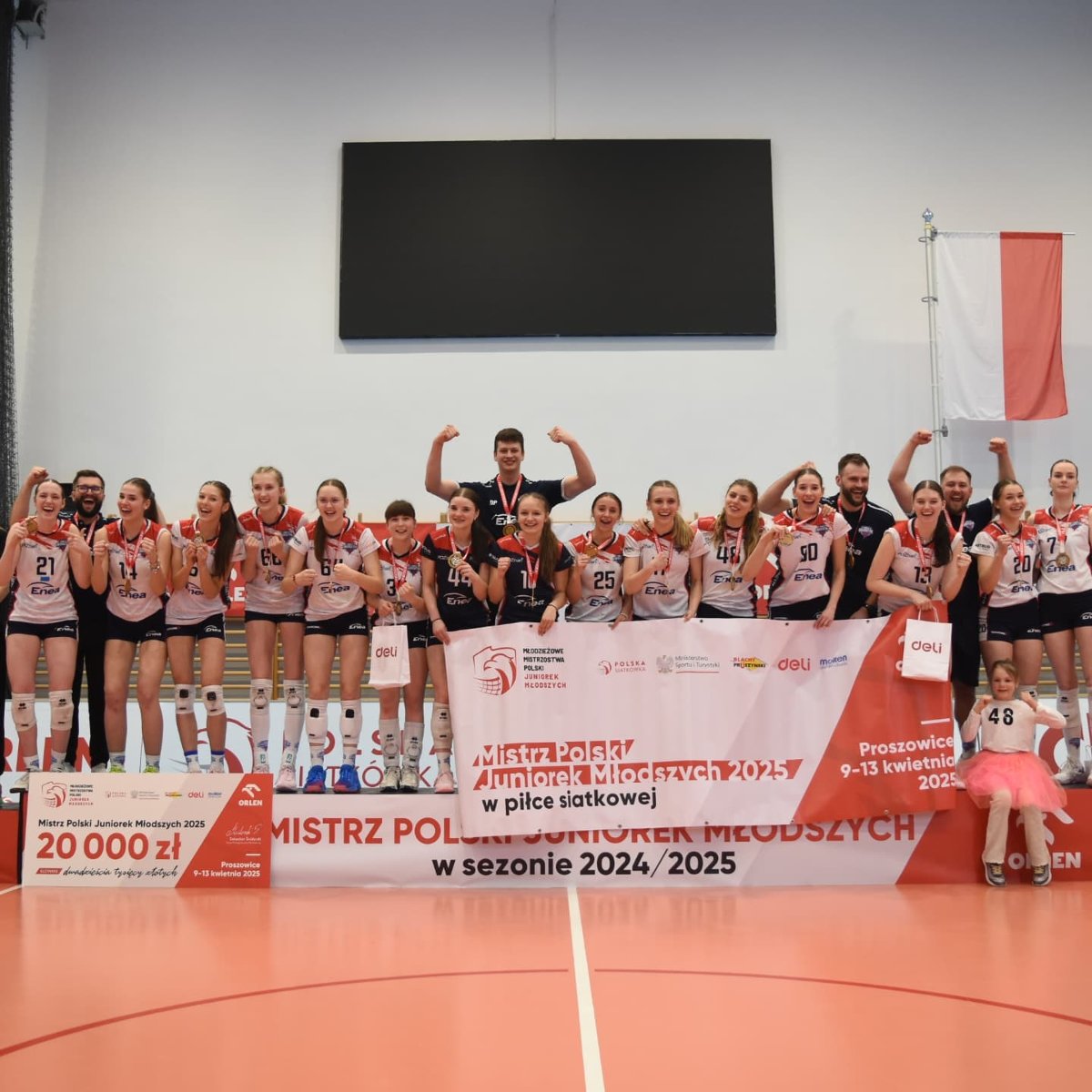 Siatkarki Enei Energetyka Poznań ze złotymi medalami Mistrzostw Polski juniorek młodszych w siatkówce, fot. Facebook Polska Siatkówka - kilkanaście zawodniczek uradowanych stoi na podium z medalami na szyi. Stoją także 4 członkowie sztabu szkoleniowego i trzymają baner mistrowski. W tle widać białą ścianę, telebim oraz polska flaga