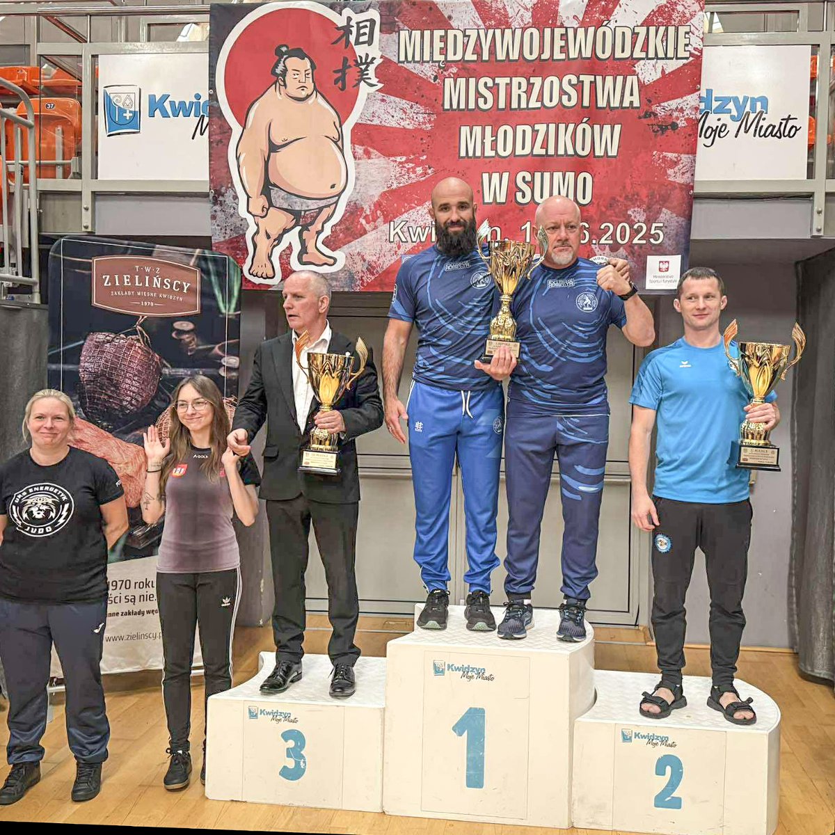 Sobieski Poznań ze zwycięstwem w klasyfikacji drużynowej kobiet podczas MMM w sumo, fot. Paulina Tim - na najwyższym stopniu podium stoi 2 mężczyzn w granatowych strojach z pucharem, na drugim i trzecim stopniu podium stoją również inni mężczyźni z pucharami, a obok podium 2 kobiety
