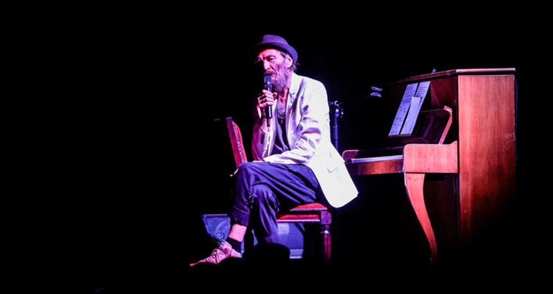 Zdjęcie ze spektaklu: przy pianinie, odwrócony do niego tyłem, na krześle siedzi mężczyzna z mikrofonem w ręce.