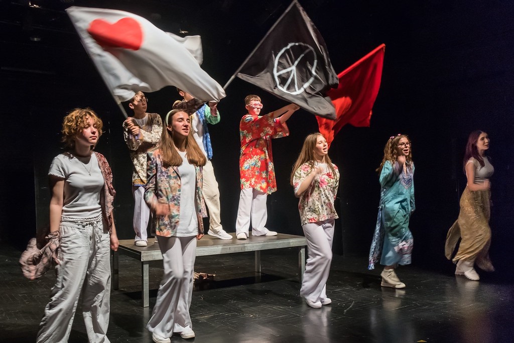 spektakl teatralny z udziałem młodych ludzi, niektórzy z nich trzymają fagi, na jednej z nich symbol pacyfizmu