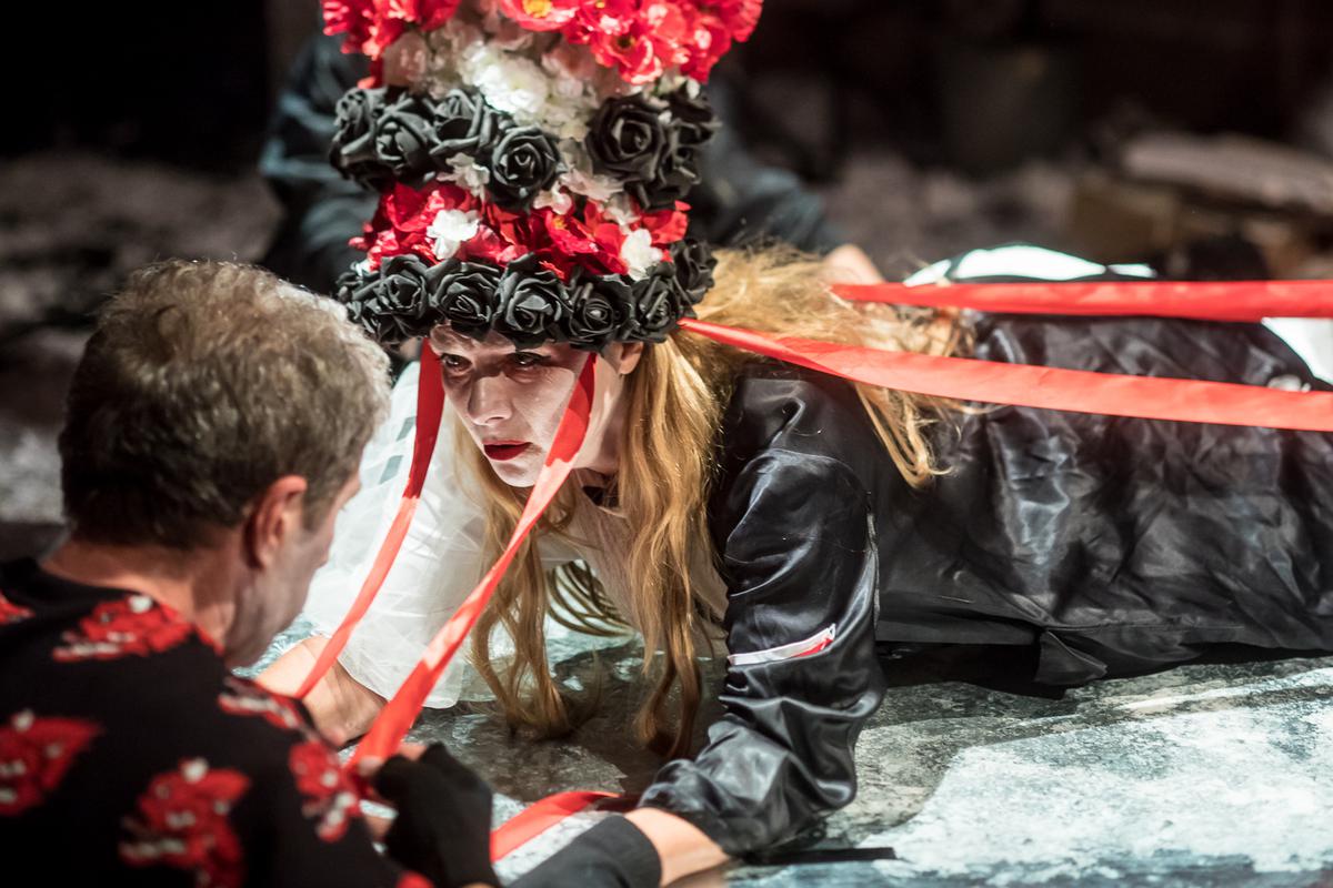 Zdjęcie ze spektaklu - kobieta z czepcem w biało-czerwono-białych kolorach na głowie, leżąca i patrzaca na mężczyznę, którego twarz jest niewidoczna.