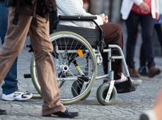Starsza osoba siedzi na wózku inwalidzkim na ulicy, widoczna jest od piersi w dół a obok niej widać nogi przechodniów