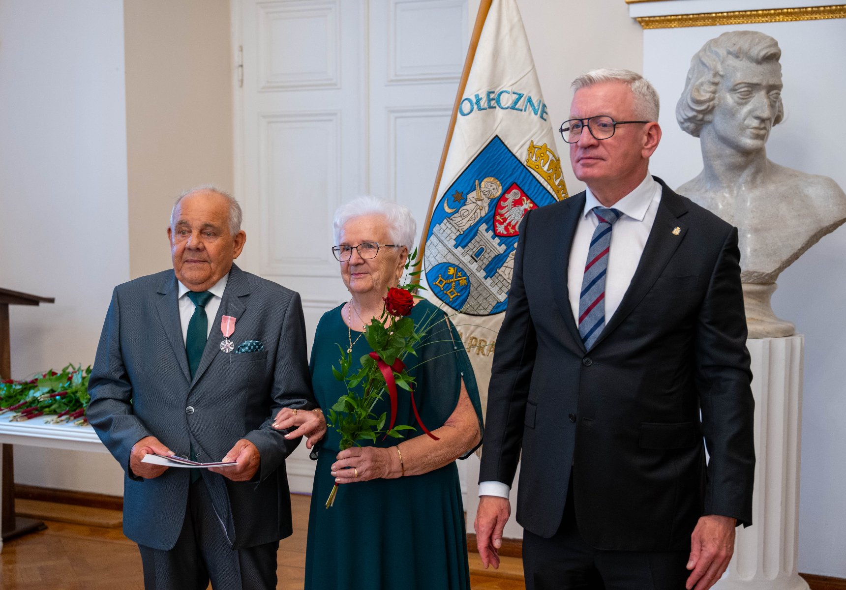 Starsza para, mężczyzna w szarym garniturze z odznaczeniem i kobieta w ciemnozielonej sukience trzymająca czerwoną różę, stoi obok mężczyzny w ciemnym garniturze i okularach.