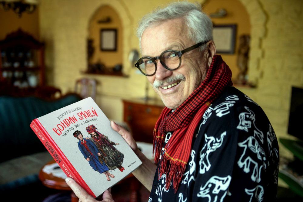 Starszy mężczyzna w szaliku pokazuje okładkę swojej książki.