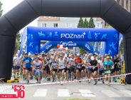 Start masowego biegu ulicznego »Poznański Czerwiec 56 - Bieg Pamięci«. Duża grupa biegaczy w kolorowych strojach sportowych przebiega pod wielką, czarną dmuchaną bramą startową. W tle widać kolejną, niebieską bramę z napisem »POZnan*« oraz logo sponsora »Enea«. Na pierwszym planie po lewej stronie znajduje się czerwono-białe logo wydarzenia z napisem »Bieg Pamięci«".