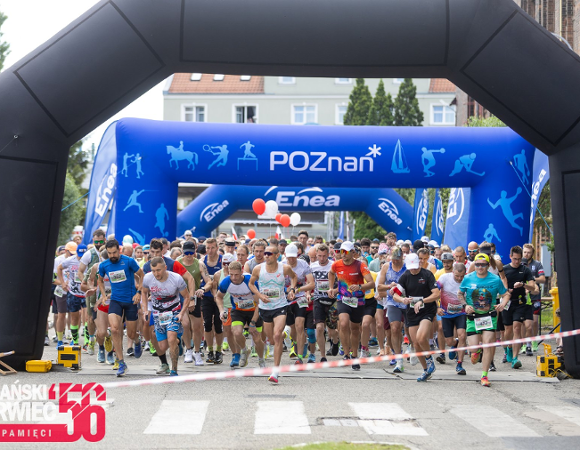 Start masowego biegu ulicznego »Poznański Czerwiec 56 - Bieg Pamięci«. Duża grupa biegaczy w kolorowych strojach sportowych przebiega pod wielką, czarną dmuchaną bramą startową. W tle widać kolejną, niebieską bramę z napisem »POZnan*« oraz logo sponsora »Enea«. Na pierwszym planie po lewej stronie znajduje się czerwono-białe logo wydarzenia z napisem »Bieg Pamięci«".