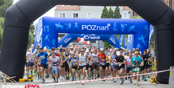 Start masowego biegu ulicznego »Poznański Czerwiec 56 - Bieg Pamięci«. Duża grupa biegaczy w kolorowych strojach sportowych przebiega pod wielką, czarną dmuchaną bramą startową. W tle widać kolejną, niebieską bramę z napisem »POZnan*« oraz logo sponsora »Enea«. Na pierwszym planie po lewej stronie znajduje się czerwono-białe logo wydarzenia z napisem »Bieg Pamięci«".