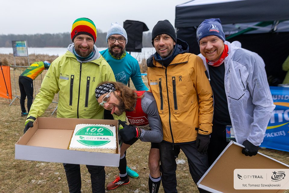 Świętowanie setnego biegu Piotra Kotkowskiego podczas serii biegów City Trail, fot. Tomasz Szwajkowski - na zdjęciu 5 mężczyzn, 3 w strojach biegowych, a 2 w zimwych. 1 z nich trzyma karton z tortem. W tle widać namiot i jezioro, a także barierki odgradzające trasę zawodów