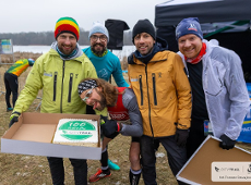 Świętowanie setnego biegu Piotra Kotkowskiego podczas serii biegów City Trail, fot. Tomasz Szwajkowski - na zdjęciu 5 mężczyzn, 3 w strojach biegowych, a 2 w zimwych. 1 z nich trzyma karton z tortem. W tle widać namiot i jezioro, a także barierki odgradzające trasę zawodów