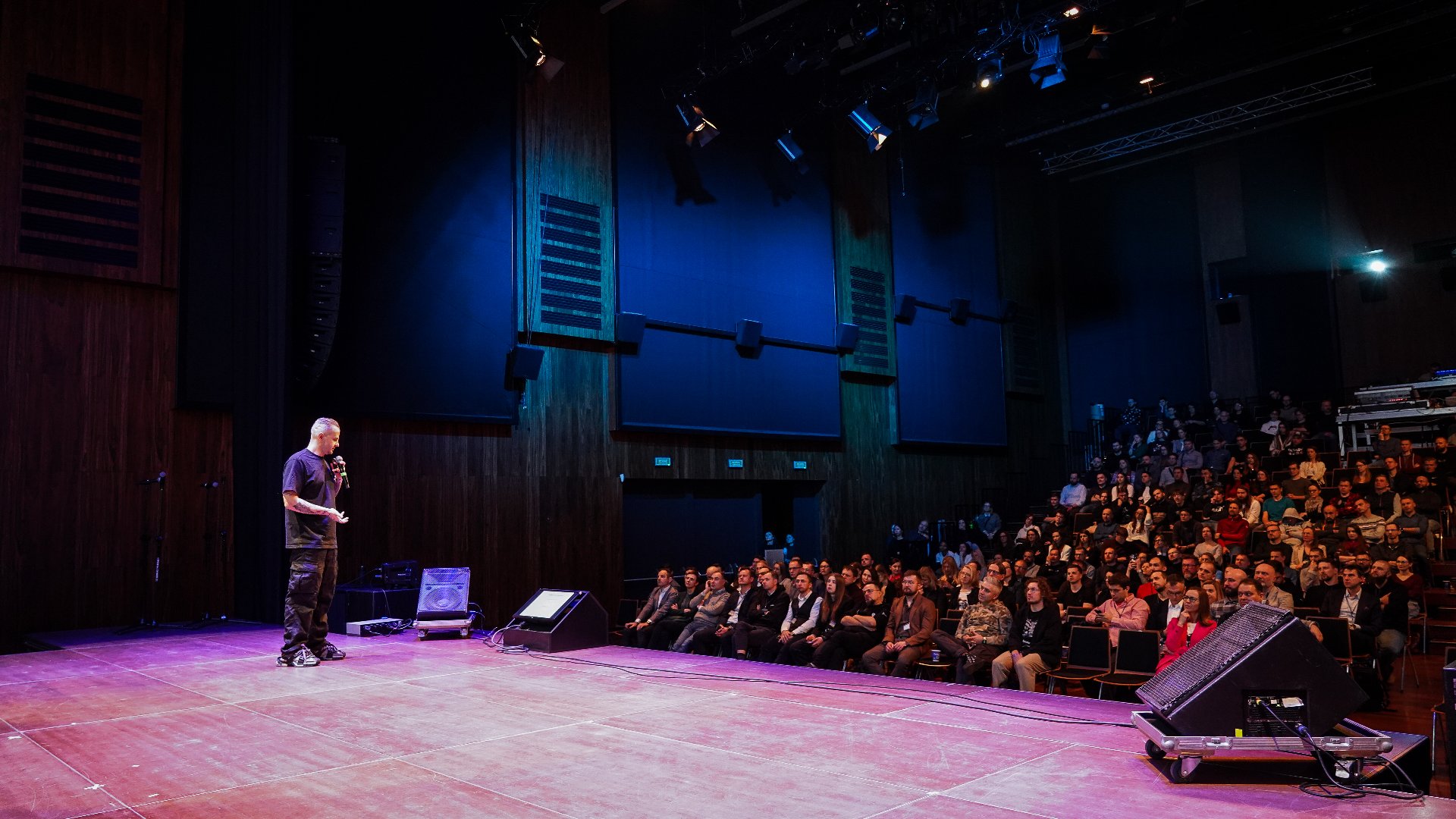 Szerokie ujęcie sali widowiskowej. Na scenie oświetlonej na czerwono stoi mężczyzna w czarnej koszulce i spodniach, trzymający mikrofon i przemawiający do publiczności. Widownia jest wypełniona słuchaczami, siedzącymi na ciemnych fotelach.